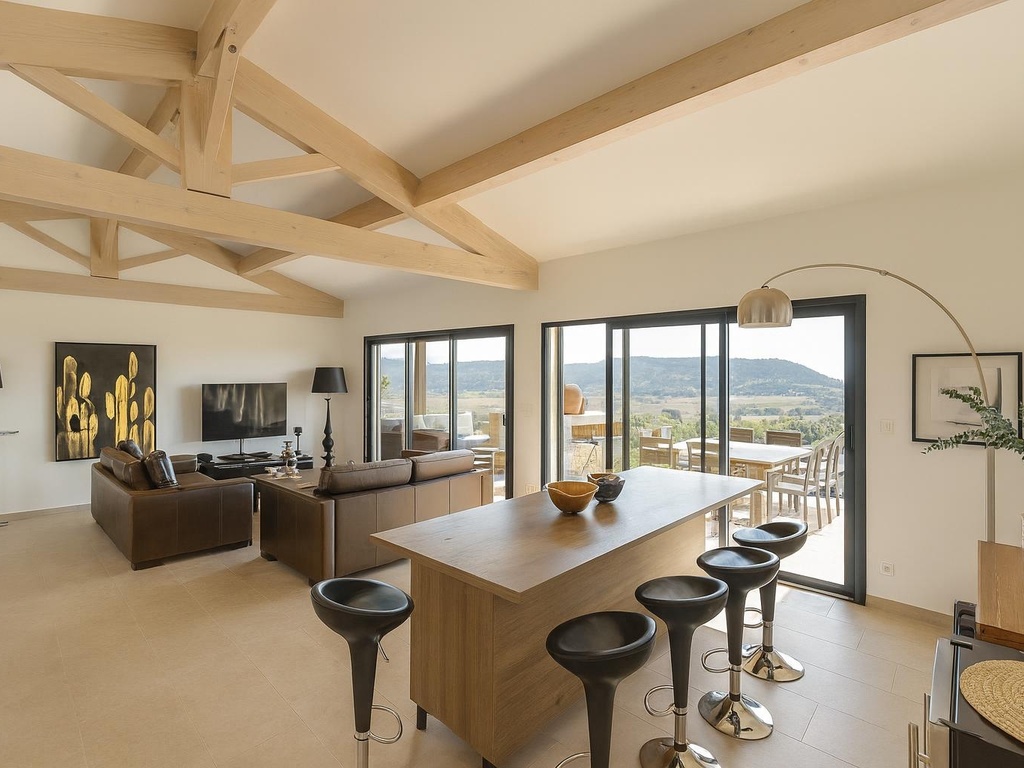 Magnifique espace de vie moderne avec cuisine ouverte, poutres apparentes, grandes baies vitrées offrant une vue panoramique sur les collines, et un aménagement contemporain avec mobilier design.