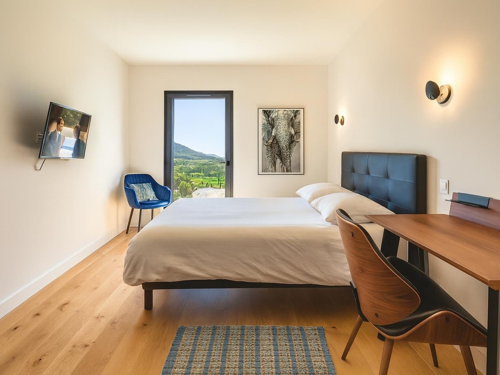 Chambre moderne et lumineuse avec lit double, télévision murale, bureau en bois, fauteuil bleu confortable et une belle vue sur les montagnes à travers une grande porte-fenêtre.