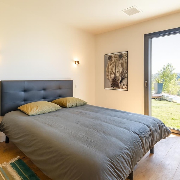 Chambre contemporaine lumineuse avec lit double, tête de lit capitonnée grise, parquet en bois clair, et grande baie vitrée offrant une vue magnifique sur la campagne.