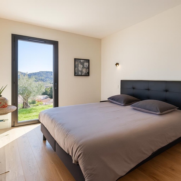 Chambre moderne et lumineuse avec lit double confortable, télévision murale, et une grande baie vitrée offrant une vue splendide sur les collines environnantes.
